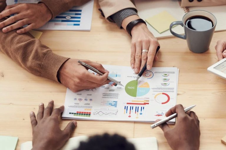 Equipe de trabalho contendo 4 pessoas em uma mesa discutindo sobre umas analises em uns papéis encima da mesa.